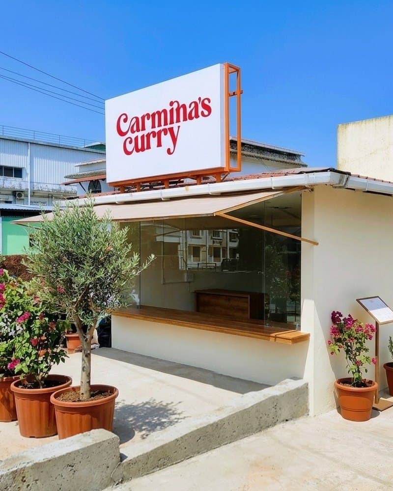 The Carmina's Curry storefront at Plot L 89, Phase 2-E, Verna Industrial Estate. Red wordmark sign on a white kitchen front, with the open glass counter and bougainvilleas at the entrance.